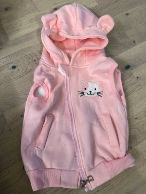 Rosa väst med kattmotiv och huva - Söt rosa väst med dragkedja, huva med öron och broderad katt på bröstet. Västen har två fickor fram och är fodrad med mjukt vitt tyg på insidan. Perfekt för dig som gillar gulliga detaljer och djurmotiv.Rosa väst med kattmotiv och huva, 2år