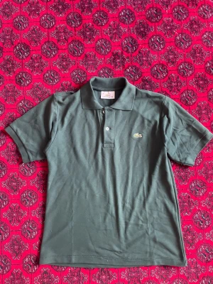 Lacoste Piké  - Klassisk grön pikétröja från Lacoste med korta ärmar och krage. Tröjan har knappar framtill och den ikoniska broderade krokodilloggan på bröstet. Perfekt för en stilren och avslappnad look.
