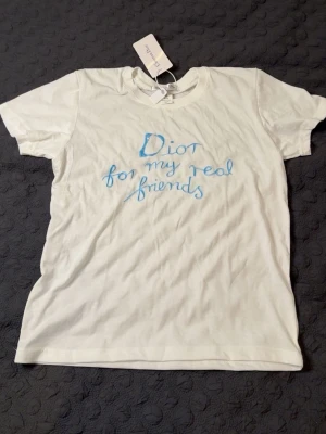 Vit CD t-shirt med blå text - Vit t-shirt från CD med handskriven blå text 'CD for my real friends' på framsidan. Klassisk rund hals och korta ärmar, tillverkad i mjuk bomull. Perfekt för dig som vill ha en cool och exklusiv look. PASSAR XS-S. HELT NY OANVÄND.