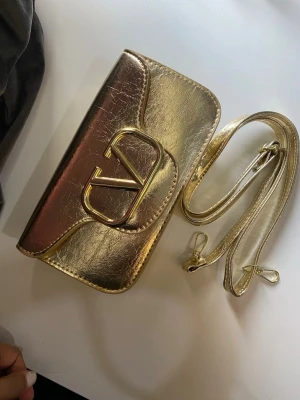 Guldig kuvertväska med axelrem - Snygg kuvertväska i metallic guld med matchande axelrem. Väskan har en stilren, rektangulär form och stängs med magnetknapp. Materialet är glansigt och ger en lyxig känsla. Perfekt för att lyfta vilken outfit som helst.