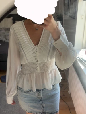 Vit blus med smock och knappar - Säljer en vit blus med v-ringning, dekorativa knappar framtill och smockad midja. Blusen har långärmade ärmar med breda manschetter och lätt volang nedtill. Materialet är tunt och luftigt. Saknas dock en knapp på varje arm (sista bilden)
