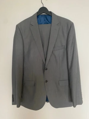 Kostym  - Snygg kostym från Dressmann i klassisk modell.Kavaj med två knappar framtill och tre fickor i storlek 50R. Fodrad med blått innerfoder och tillverkad i en mix av polyester och viskos. Perfekt för dig som vill ha en stilren och enkel look.Byxor i storlek 33/32