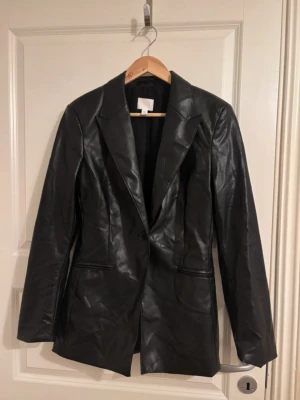 Svart fusk skinn kavaj - Snygg svart fuskskinn kavaj med klassisk krage och två fickor framtill. Jackan har en knappstängning och figurnära passform som ger en stilren look. Perfekt för dig som vill ha en trendig och tidlös jacka i garderoben. Kolla min profil för fler plagg! 🥰