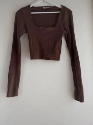 Brun croppad långärmad topp - Snygg brun croppad topp med lång ärm och fyrkantig ringning. Ribbad struktur som ger en trendig look. Perfekt att styla med höga jeans eller kjol.