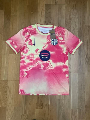 Barcelona matchtröja rosa Nike - Säljer en Barcelona matchtröja från Nike i rosa och ljusgult med molnliknande mönster. Tröjan har korta ärmar, klubbmärke och Nike-logga på bröstet samt Spotify-logga. På ryggen står det 'BALDE' och nummer 3 i blått. Perfekt för dig som älskar fotboll och unik stil.