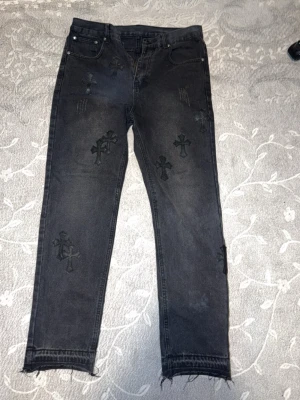 Svarta jeans med korsdetaljer - Unika svarta jeans med flera broderade kors i svart på benen. Jeansen har råa, fransiga benslut och slitna detaljer för en edgy look. Klassisk femficksmodell med dragkedja och knapp i metall. Perfekta för dig som gillar streetwear och vill sticka ut.
