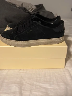 Svarta sneakers från Axel Arigato - Stilrena svarta sneakers från Axel Arigato med lågt skaft och klassisk siluett. Ovandelen är i svart mocka med diskret guldfärgad logga på sidan. Skorna har vita, något slitna sulor och svarta skosnören. Perfekta för en clean och modern look. Påse och sko låda och dust ag kommer med. 