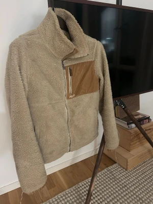 Beige teddyjacka från Polo Ralph Lauren - Säljer en beige teddyjacka från Polo Ralph Lauren med dragkedja framtill och hög krage. Jackan har en snygg detalj i form av en bröstficka i brunt manchestertyg med dragkedja. 
