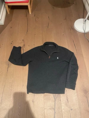 Mörkgrå halvzip från Polo Ralph Lauren - Mörkgrå tröja med halv dragkedja från Polo Ralph Lauren. Klassisk broderad logga i vitt på bröstet och läderdetalj på dragkedjan. Tröjan har lång ärm och en avslappnad passform, perfekt för en stilren och enkel look.