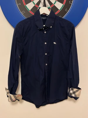 Burberry Skjorta mörkblå  - Snygg mörkblå skjorta från Burberry med klassiskt rutigt foder i ärmsluten och liten logga på bröstet. Skjortan har knappar framtill och lång ärm. Perfekt för dig som gillar stilrena plagg med exklusiva detaljer. Knapp på ena armen saknas men har ingen större betydelse, går att ersätta enkelt.