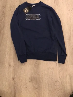 Mörkblå sweatshirt från MUSIC - Mörkblå sweatshirt från MUSIC med trycket 'Original Street Style New York City' på bröstet. Tröjan har rund hals, ribbade muddar vid ärmslut och nederkant samt en avslappnad passform. Perfekt för en casual look.