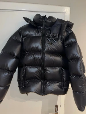 Svart pufferjacka från J.Lindeberg - Säljer en svart pufferjacka från J.Lindeberg med glansig finish och huva. Helt ny, aldrig använd. Jackan har dragkedja framtill, två sidofickor med dragkedja och ett stilrent JL-logo på bröstet. Perfekt för de kalla vinterdagarna. Tags medföljer ej. Skickas samma dag. Nypris runt 4500-5500kr. Pris kan diskuteras. Hör av er vid intresse. 