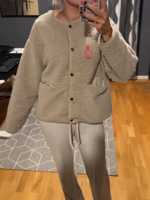 Beige teddyjacka med knappar - Mysig beige teddyjacka med rund halsringning och svarta tryckknappar framtill. Jackan har två fickor och en liten rosa detalj på bröstet. Oversized passform och fluffigt material som ger en trendig look.