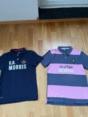 Randig pikétröja från Morris - Säljer en klassisk pikétröja från Morris med breda rosa och mörkblå ränder. Tröjan har krage, knappar framtill och broderad logga på bröstet. Texten 'Isle of Wight MORRIS' är tryckt på framsidan. Perfekt för en sportig och avslappnad stil.