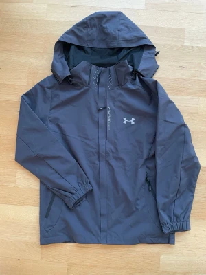 Svart vindjacka från Under Armour - Snygg svart vindjacka från Under Armour med huva och dragkedja framtill. Jackan har två sidofickor med dragkedja, elastiska muddar vid ärmsluten och meshfoder för extra komfort. Perfekt för blåsiga dagar och sportiga outfits.