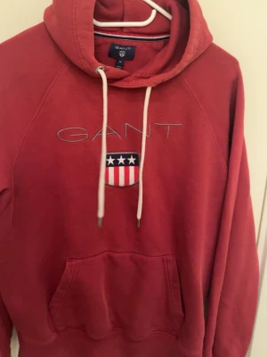 Röd hoodie från GANT med brodyr - Röd hoodie från GANT med broderad logga och amerikanskinspirerat sköldmärke på bröstet. Klassisk känguruficka framtill, vita dragsnören och huva. Mjuk bomullsblandning och avslappnad passform, perfekt för en casual look.
