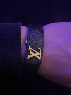 Louis Vuitton svart läderarmband - Snyggt svart armband från Louis Vuitton i präglat läder med det ikoniska LV-emblemet i guldmetall framtill. Armbandet har en bred design och stängs med ett metallspänne. Perfekt accessoar för dig som gillar exklusiv och trendig stil.  📦FRAKTAR SAMMA DAG SOM KÖP!