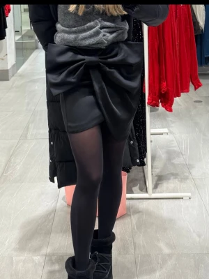 Svart minikjol med stor rosett - Supersnygg svart minikjol i satinliknande material med en stor, statement rosett bak. Kjolen har en tight passform och är perfekt för dig som vill sticka ut. Perfekt att styla med strumpbyxor och boots.