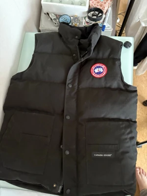 Svart dunväst från Canada Goose - Snygg svart dunväst från Canada Goose med klassisk logga på bröstet och två stora fickor framtill. Västen har hög krage och stängs med både dragkedja och knappar. Perfekt för lager-på-lager och håller dig varm under kyliga dagar.