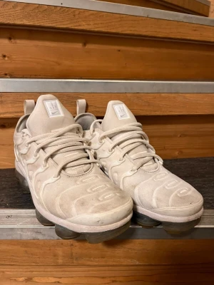 Nike Air Vapormax Plus vita sneakers - Nike Air Vapormax Plus sneakers i vitt med ikonisk plastbur på ovandelen och genomskinlig sula med synliga luftbubblor. Skorna har en sportig siluett, rund tå och klassiska Nike-detaljer på plösen. Perfekta för dig som gillar streetwear och vill ha en unik look.