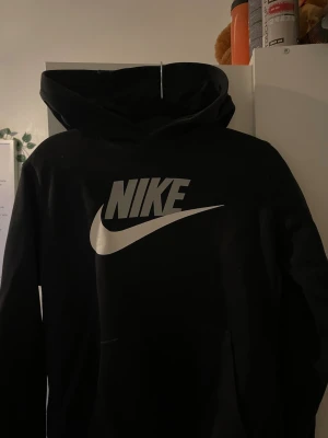 Svart Nike hoodie med stor logga - Svart hoodie från Nike med stor vit logga och swoosh på bröstet. Klassisk standard fit med huva och känguruficka framtill. Perfekt för en sportig och avslappnad stil. Tillverkad i mjukt material för extra komfort.