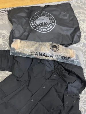 Svart dunjacka från Canada Goose - Säljer en svart dunjacka från Canada Goose med avtagbar huva och pälsdetalj. Jackan har tryckknappar, dragkedja och två stora fickor framtill. Klassisk patch på ärmen och medföljande förvaringspåse. Perfekt för kalla vinterdagar. Jackan har två hål som man kan se på bilderna och de är sydda.