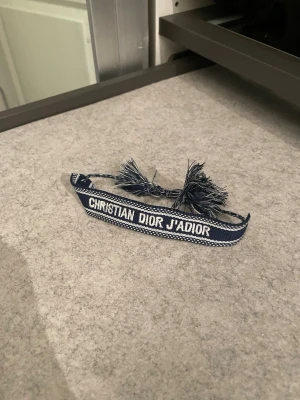 Christian Dior armband - Snyggt blått tygarmband från Christian Dior med texten 'CHRISTIAN DIOR J'ADIOR' broderad i vitt. Armbandet har dekorativa tofsar i ändarna och justerbar knytning. Perfekt accessoar för att lyfta din outfit. 🔥Bara höra av sig i dm vid frågor och funderingar!🔥
