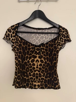 Leopardmönstrad mesh topp med v-ringning - Trendig leopardmönstrad topp i mesh med v-ringning och svarta spetsdetaljer längs halsen. Kortärmad och croppad modell som ger en edgy look. Perfekt för dig som vill sticka ut med ett djurmönster.