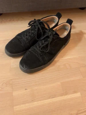 Svarta sneakers med röd sula - Snygga svarta sneakers från Christian Louboutin med klassisk röd sula. Skorna har lågt skaft, svarta skosnören och ovandel i mocka. Perfekta för dig som vill sticka ut med en ikonisk detalj under foten.
