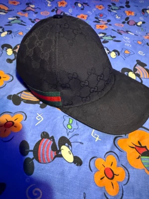 Svart Gucci keps med GG-mönster - Säljer en svart keps från Gucci med det klassiska GG-monogrammet över hela kepsen. På sidan finns en grön och röd rand som ger extra Gucci-vibe. Kepsen har böjd skärm och är tillverkad i ett slitstarkt tygmaterial. Perfekt för dig som vill ha en stilren och lyxig accessoar. Rensning sker hos brorsan. 