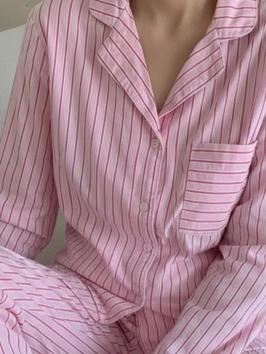 Rosa randig pyjamas med skjorta - Säljer ett set med pyjamas i mjuk bomull, ljusrosa med tunna röda ränder. Överdelen har klassisk skjortkrage, knäppning framtill och bröstficka. Byxorna har resår i midjan för extra komfort. Perfekt för mysiga kvällar hemma.