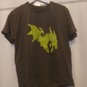 Grön t-shirt med neonmönster, M - Säljer en grön Närcon t-shirt från 2024  i storlek M med ett stort, pixlat neon-grönt motiv på framsidan. T-shirten har klassisk rund hals och korta ärmar. Materialet känns som mjuk bomull, perfekt för dig som gillar grafiska tryck och vill sticka ut.