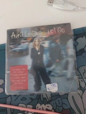 Avril Lavigne Let Go CD-album - Säljer Avril Lavignes ikoniska album 'Let Go' på CD. Omslaget är i färg med artistens namn och albumtitel, och baksidan visar låtlistan. Albumet innehåller hits som 'Complicated' och 'Sk8er Boi'. Perfekt för dig som gillar poprock och samlar på CD-skivor.