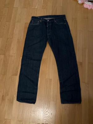 Mörkblå raka jeans från Levi's - Klassiska mörkblå jeans från Levi's med rak passform och fem fickor. Jeansen har tydliga kontrastsömmar och en läderpatch bak i midjan. Perfekta för en stilren och tidlös look.