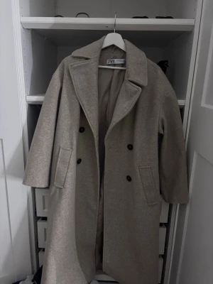 Beige ullkappa från Zara - Stilren beige ullkappa från Zara med dubbelknäppning och breda slag. Kappan har två stora fickor framtill