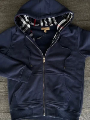 Marinblå hoodie från Burberry - Snygg marinblå hoodie från Burberry med dragkedja och klassiskt rutigt foder i huvan. Hoodien har dragsko vid huvan, två fickor framtill och ribbade muddar. Perfekt för dig som gillar stilrena och exklusiva plagg med en twist.