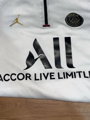 PSG vit matchtröja Jordan - Säljer en vit PSG matchtröja från Jordan med svarta detaljer och guldigt Jumpman-märke. Tröjan har svart Paris Saint-Germain-logga på bröstet och vinröda inslag på ärmarna. Tillverkad i lätt och ventilerande material, perfekt för fotboll eller träning.