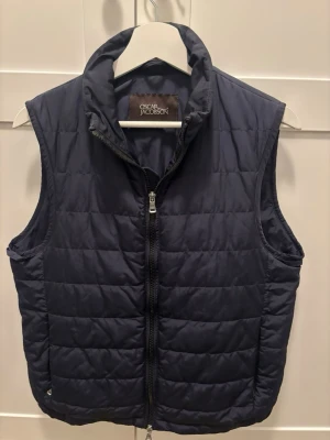 Svart dunväst från Oscar Jacobson - Säljer en marinblå dunväst från Oscar Jacobson med hög krage och hel dragkedja framtill. Västen har en stilren och enkel design, perfekt att slänga över en hoodie eller tröja. Materialet är mjukt och lätt, vilket gör den smidig att bära under kyligare dagar.