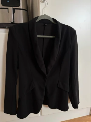 Svart kavaj  - Stilren svart kavaj med en knapp framtill. Kavajen har två fickor med lock och är figursydd för en snygg siluett. Perfekt att slänga över en t-shirt eller skjorta för en clean look.