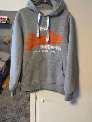 Grå Superdry hoodie med tryck - Snygg grå hoodie från Superdry med stor orange och vit logga på bröstet samt japansk text. Inga defekter förutom att trycket är lite slitet 