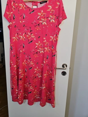 Rosa blommig klänning från Signature - Snygg rosa klänning från Signature med blommigt mönster i orange och svart. Modellen är kortärmad och har en rund halsringning. Perfekt för dig som gillar färg och mönster. Material 95% viskos 5% elastan 