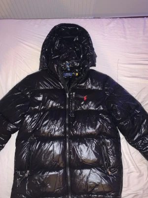Svart dunjacka från Polo Ralph Lauren - Snygg svart dunjacka från Polo Ralph Lauren med blank finish och röd broderad logga på bröstet. Jackan har huva, dragkedja framtill och två sidofickor med dragkedja. Perfekt för kalla vinterdagar.