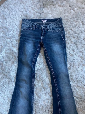 Blå bootcut jeans med broderade fickor - Säljer ett par blå bootcut jeans med snygga slitningar och vita kontrastsömmar. Baksidan har två fickor med stora broderade mönster i vitt och grått. Jeansen har klassisk femficksmodell och normal midja. Perfekta för dig som gillar detaljer!