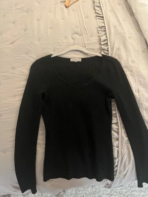 Svart ribbad tröja med v-ringning - Snygg svart ribbad tröja med lång ärm och djup v-ringning. Figurnära passform och stretchigt material som sitter skönt på kroppen. Perfekt basplagg till garderoben.