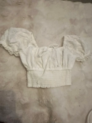 Vit broderad blus med puffärm - Supersöt vit blus med broderade detaljer och puffiga korta ärmar. Toppen har smockad midja och knyt framtill för extra söt look. Perfekt till höga jeans eller kjol för en trendig outfit.