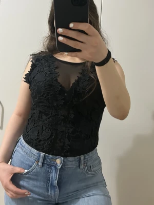 Svart spetslinnetopp med meshdetaljer - Svart spetsbody i mycket fint skick, oanvänd. Endast testad, säljs då den inte kommer till användning. Sitter fint och är stretchig. Genomskinlig i ryggen.  Storlek M, passar även S. Pris kan diskuteras 💗