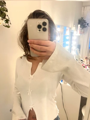 Vit ribbad topp med rosetter från Cospard - Supersöt vit ribbad topp från Cospard med lång ärm och tre små rosetter framtill. Toppen har en lätt croppad passform och v-ringad hals. Perfekt att styla med jeans eller kjol för en trendig look.