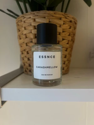 ESSNCE Carashmellow Eau de Parfum - Minimalistisk parfymflaska från ESSNCE med doften Carashmellow. Flaskan är cylindrisk i genomskinligt glas med svart rund kork och enkel vit etikett med svart text. Perfekt för dig som gillar stilrena och moderna doftflaskor.
