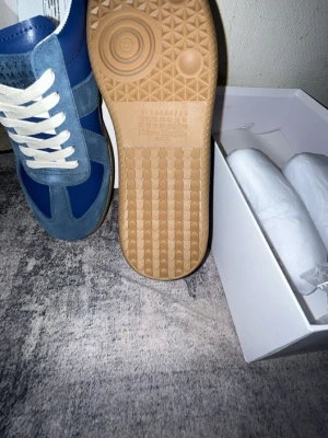 Blå sneakers från Maison Margiela - Snygga blå sneakers från Maison Margiela med vita skosnören och detaljer i ljusblå mocka. Skorna har en klassisk låg siluett och en brun gummisula med unikt mönster undertill. Perfekta för dig som gillar stilrena och trendiga sneakers.