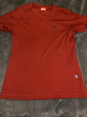 Röd funktions-topp från Fjällräven - Snygg röd funktions-topp från Fjällräven med V-ringning och diskret logga på bröstet. Storlek xs och nästan helt oanvänd. 